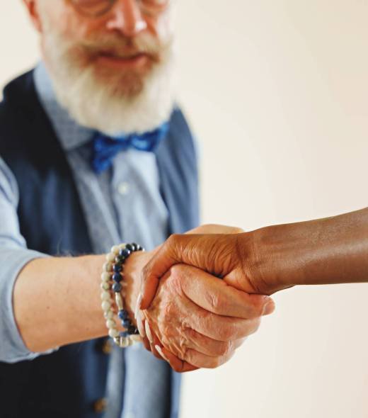 Eccentric, stylish man with a beard and mustache shaking hands with an African woman. The handshake is the focus, the man's face is blurred, and the background is neutral.