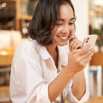 Vertical shot of brunette asian girl sitting in cafe, using smartphone app, chatting on mobile phone and smiling.